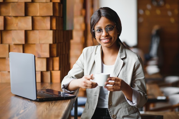 Ritratto di una donna afroamericana sorridente felice seduta al tavolo in un bar, guardando la fotocamera, eccitata in posa femminile, lavorando al computer, facendo i compiti, preparando un rapporto in una caffetteria