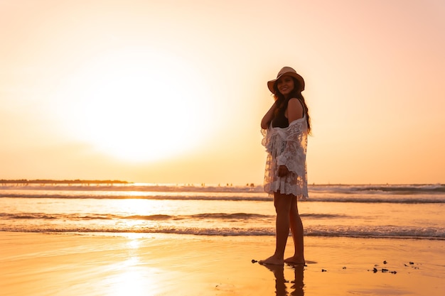 Ritratto di una donna al tramonto in un abito bianco con un cappello che cammina in riva al mare con la bassa marea