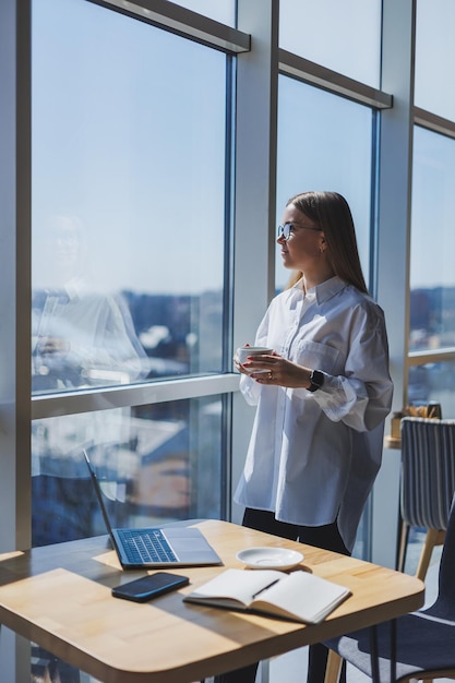 Ritratto di una donna allegra con gli occhiali classici che sorride nel suo tempo libero in un bar con caffè donna ebrea positiva in una camicia bianca scrivania con computer portatile lavoro a distanza