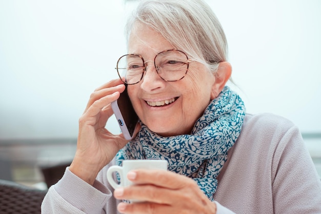 Ritratto di una donna anziana sorridente seduta all'aperto con una tazza di caffè espresso che parla con lo smartphone