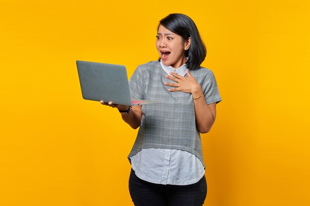 Ritratto di una donna asiatica eccitata che riceve e-mail in arrivo sul laptop su sfondo giallo