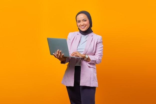 Ritratto di una donna asiatica sorridente che tiene in mano un laptop e guarda la telecamera su sfondo giallo