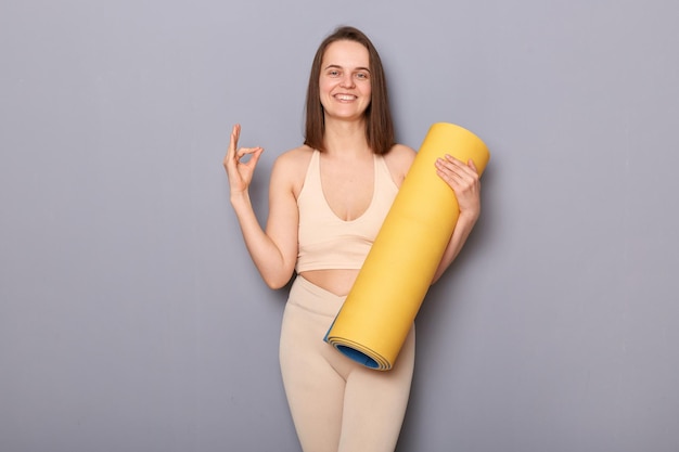 Ritratto di una donna atletica piuttosto positiva che tiene il tappetino fitness arrotolato e mostra il segno giusto approvare la nuova palestra e l'istruttore che si gode l'allenamento maditando guardando sorridente alla macchina fotografica