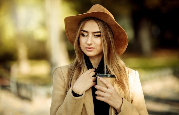 Ritratto di una donna attraente con il trucco vestito con un cappotto e un cappello di feltro. Passeggia nel parco cittadino autunnale