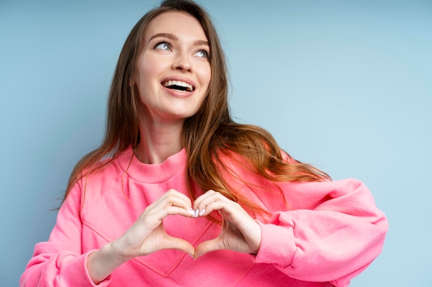 Ritratto di una donna attraente e sorridente che indossa una felpa rosa con un sorriso dentato che fa un gesto al cuore