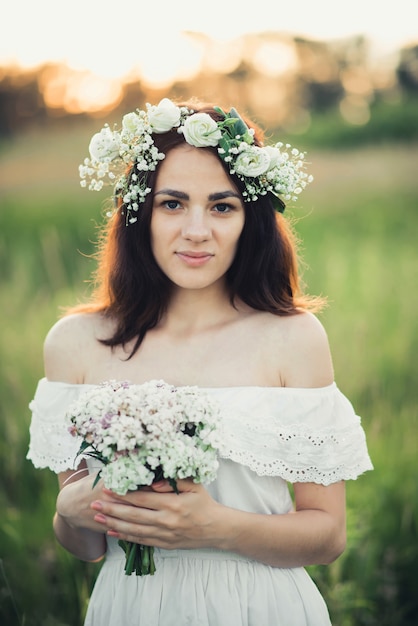 Ritratto di una donna attraente in abito bianco con un mazzo di fiori e una corona in estate