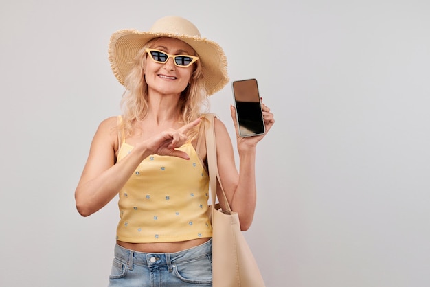 Ritratto di una donna bionda matura con cappello di paglia, top giallo e occhiali da sole che tiene uno smartphone e una borsa da spesa isolata sullo sfondo bianco dello studio