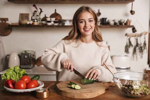 ritratto di una donna carina e contenta che indossa abiti casual sorridenti e prepara il pranzo in una cucina accogliente