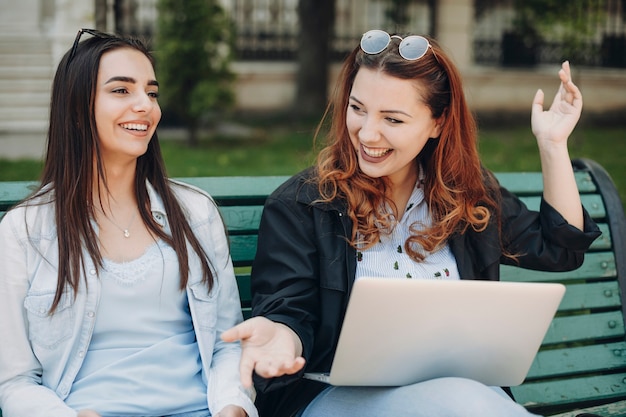 Ritratto di una donna caucasica adorabile due che si siede sulla panchina che ride mentre tiene un computer portatile sulle gambe.