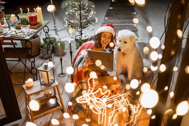 Ritratto di una donna con il suo simpatico cane che celebra le vacanze di Capodanno sulla terrazza splendidamente decorata di casa, con una videochiamata su un tablet digitale