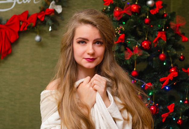 Ritratto di una donna con labbra rosse luminose, capelli lunghi biondi contro l'albero di Capodanno. Giovane donna in un cappotto bianco caldo, le spalle sono visibili. Vacanze. buon Natale