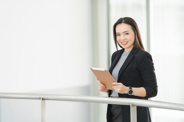 Ritratto di una donna d'affari asiatica allegra e fiduciosa in giacca e cravatta in piedi mentre usa una tavoletta digitale nell'edificio aziendale. Foto d'archivio aziendale