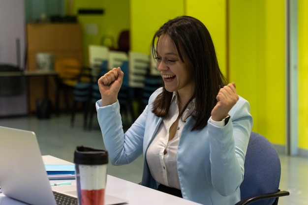Ritratto di una donna d'affari con un gesto celebrativo con le braccia in su per il successo nella sua azienda seduta davanti al computer nel suo ufficio al lavoro