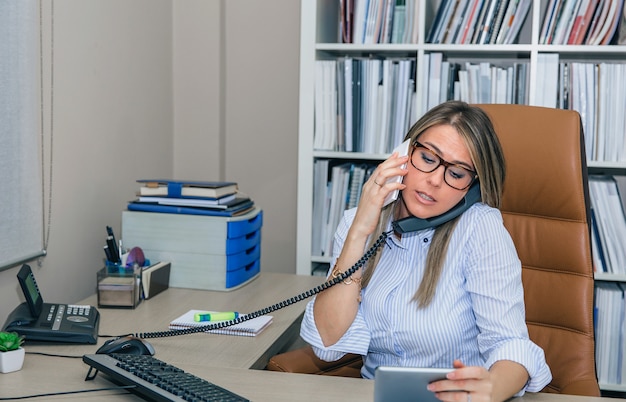 Ritratto di una donna d'affari impegnata che parla contemporaneamente al cellulare e al telefono fisso mentre guarda un tablet elettronico nelle sue mani