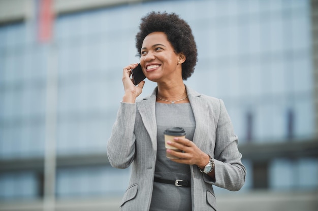 Ritratto di una donna d'affari nera di successo che utilizza l'app su uno smartphone mentre fa una pausa caffè davanti a un edificio aziendale.
