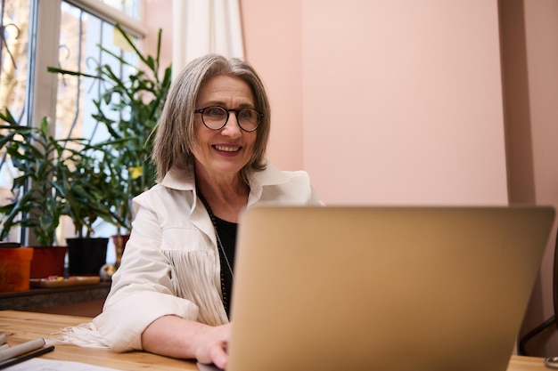 Ritratto di una donna europea anziana piacevole che sorride mentre lavora al computer portatile nel suo ufficio