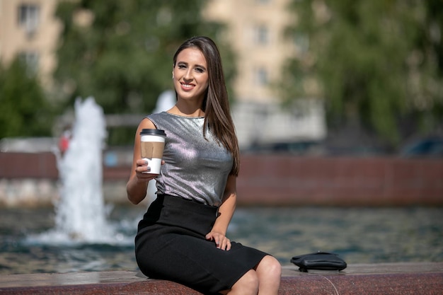 Ritratto di una donna europea sorridente con una maglietta grigia lucida che passeggia per le strade della città con caffè da asporto in mano