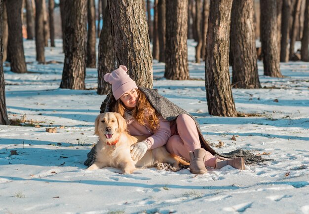 Ritratto di una donna felice che abbraccia il suo cane golden retriever coperto insieme con un plaid su un bosco innevato.