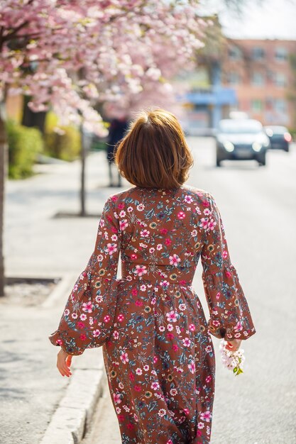 Ritratto di una donna gentile sullo sfondo di fiori di sakura. Passeggia nel giardino fiorito di sakura. Giovane donna alla moda che sta nel parco di sakura e che gode della bellezza