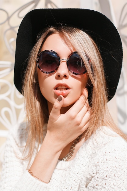 Ritratto di una donna giovane e bella hipster in un elegante cappello nero in occhiali da sole rotondi in un maglione bianco lavorato a maglia in una soleggiata giornata estiva. Ragazza alla moda sexy.