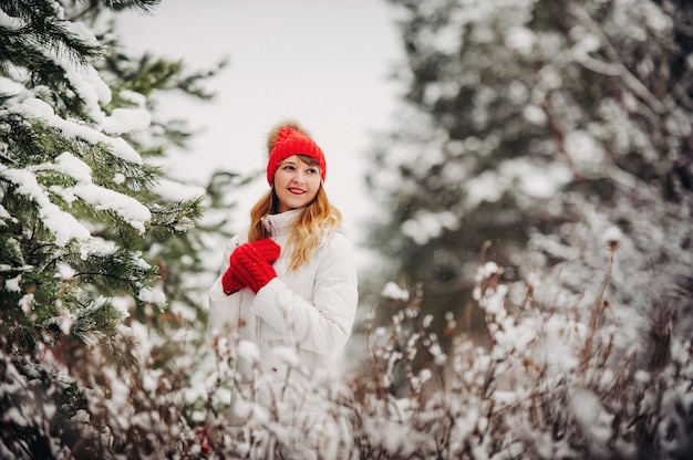 Ritratto di una donna in abiti bianchi e un cappello rosso in una fredda foresta invernale. Ragazza in una foresta invernale innevata