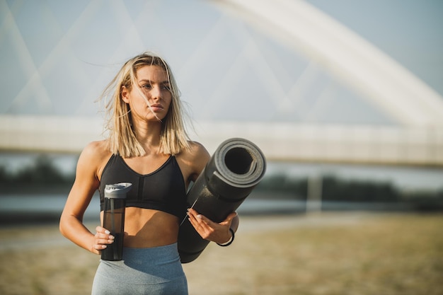 Ritratto di una donna in forma in abbigliamento sportivo che tiene un tappetino da yoga e una bottiglia d'acqua e si prepara per l'allenamento vicino al fiume, in città.