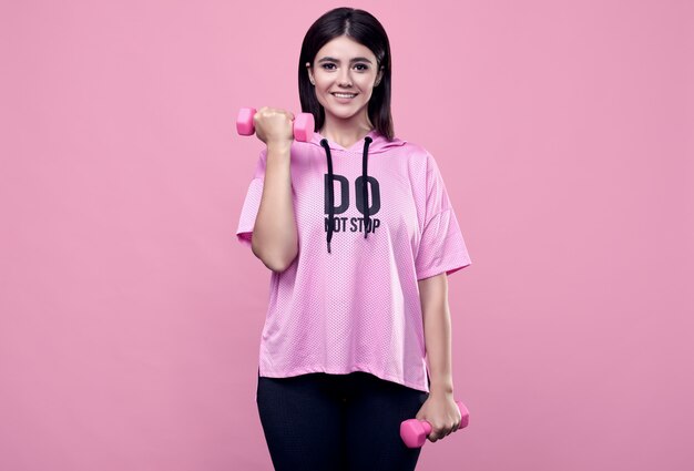 Ritratto di una donna latina positiva del corpo splendido in una felpa con cappuccio rosa di sport che si esercita con le teste di legno sul rosa