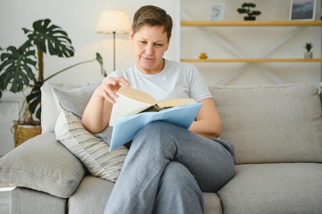 Ritratto di una donna matura felice che si rilassa sul divano e legge un libro in una casa moderna