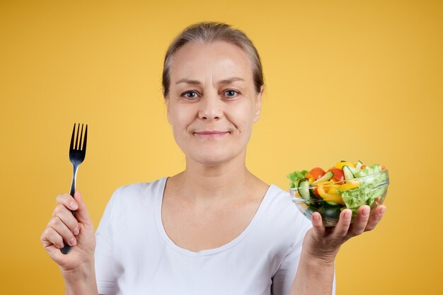 Ritratto di una donna matura sorridente in una camicia bianca che tiene un piatto di insalata e una forchetta, pronta per la cena