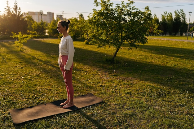 Ritratto di una donna pacifica che indossa abiti sportivi bianchi in piedi nel parco della città, godendo di un momento di consapevolezza senza stress, facendo esercizi di yoga, rilassamento, occhi chiusi, meditando sulla natura la sera durante il tramonto