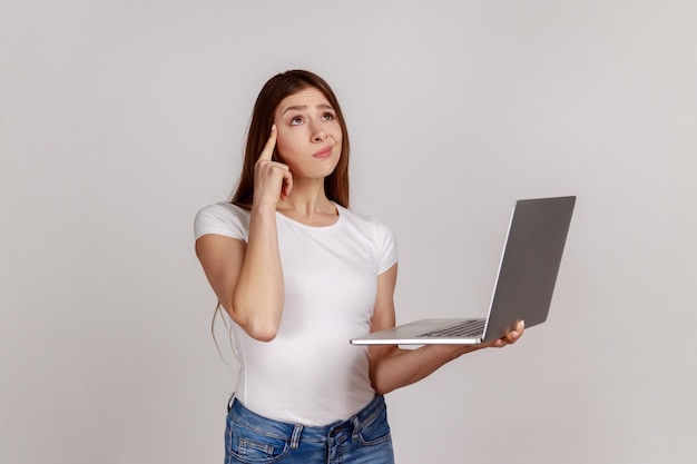 Ritratto di una donna premurosa e confusa che tiene il laptop pensando a una domanda difficile, riflettendo sulla soluzione, indossando una maglietta bianca. Studio indoor girato isolato su sfondo grigio.