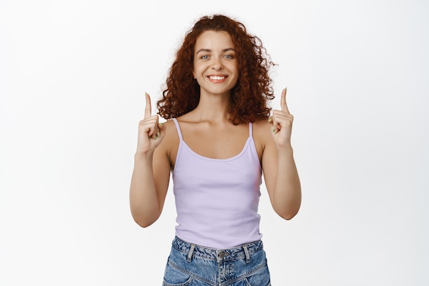 Ritratto di una donna rossa dai capelli ricci sorridente, che sembra felice, che punta le dita in piedi su bianco