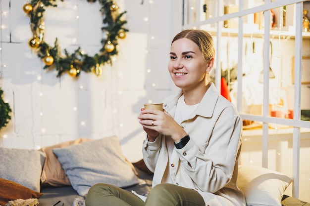 Ritratto di una donna sorridente con caffè profumato tra le mani sullo sfondo delle decorazioni di Capodanno Caffè festivo di Natale a casa sul letto Vacanze di Capodanno