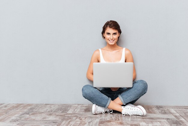 Ritratto di una donna sorridente seduta sul pavimento con un computer portatile isolato su sfondo grigio