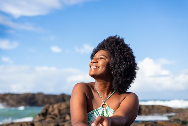 Ritratto di una donna sorridente vestita di blu seduta sulla roccia di una spiaggia guardando di lato Cielo blu sullo sfondo Spiaggia di Rio Vermelho Salvador Brasile