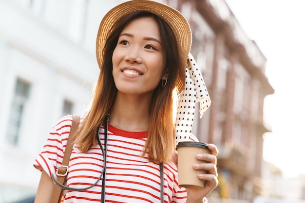 Ritratto di una donna turistica asiatica soddisfatta che indossa un cappello di paglia che beve caffè da asporto mentre cammina per la strada della città