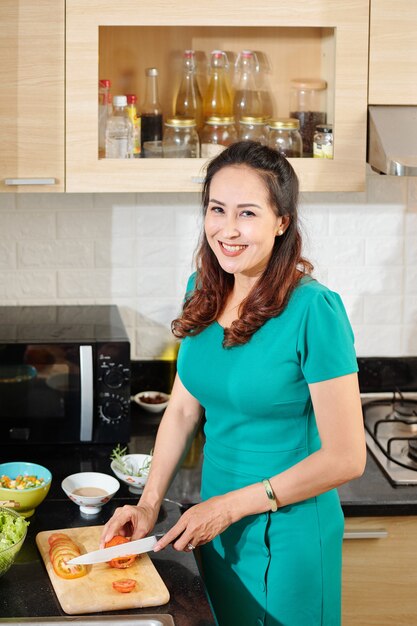 Ritratto di una donna vietnamita di mezza età positiva che taglia i pomodori per una sana insalata per cena