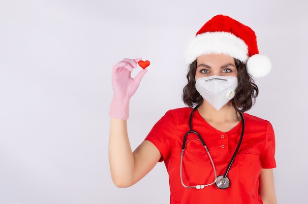 Ritratto di una dottoressa in una maschera protettiva medica Cappello di Babbo Natale e guanti in nitrile rosa mano con l'immagine di un cuore un simbolo di amore cardiologia