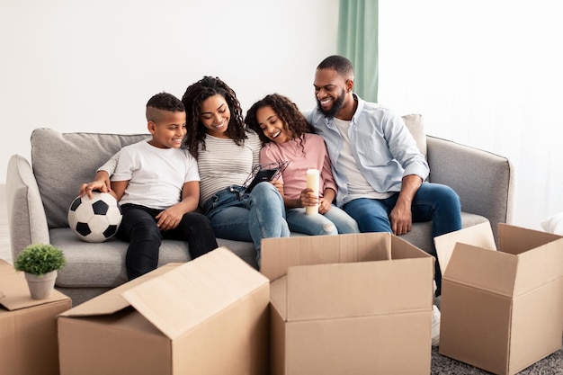 Ritratto di una famiglia nera sorridente di quattro persone sedute sul divano in soggiorno, che si trasferiscono o si trasferiscono, disimballano scatole di cartone, tengono la cornice e guardano insieme la loro foto