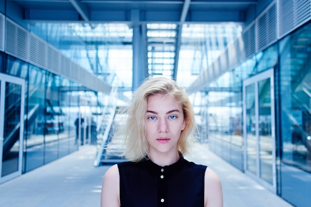 Ritratto di una forte ragazza bionda in nero dal futuro sullo sfondo di un edificio commerciale di vetro guardando la telecamera