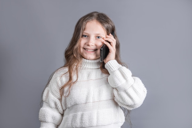 Ritratto di una giovane bambina attraente con capelli fluenti lunghi biondi in un maglione bianco, parlando al telefono