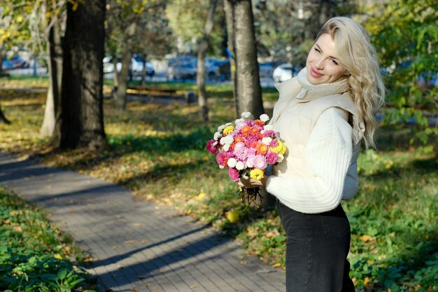 Ritratto di una giovane bella bionda con i capelli lunghi in un maglione bianco e giacca in un soleggiato autunno in un parco della città con un bouquet di fiori