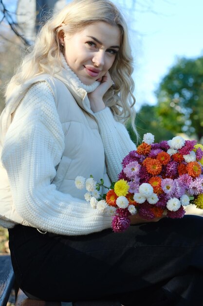 Ritratto di una giovane bella bionda con i capelli lunghi in un maglione bianco e giacca in un soleggiato autunno in un parco della città con un bouquet di fiori
