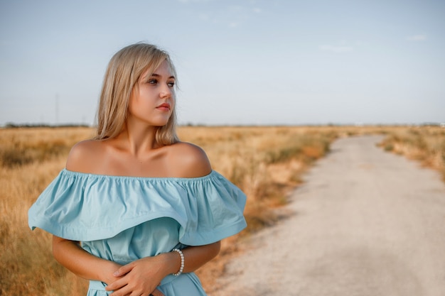 Ritratto di una giovane bella ragazza bionda caucasica in un abito azzurro in piedi su un campo con erba seccata al sole accanto a una piccola strada di campagna