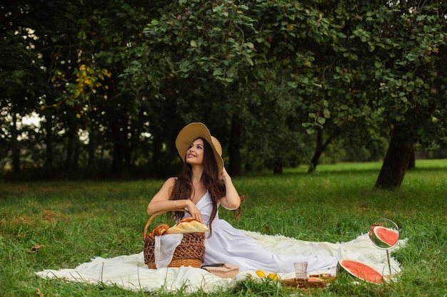 Ritratto di una giovane bella ragazza con denti anche bianchi, un bel sorriso in un cappello di paglia e un lungo abito bianco fanno un picnic in giardino.