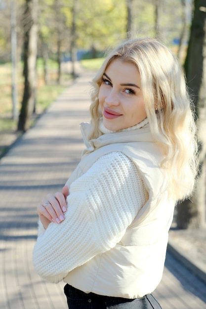 Ritratto di una giovane bellissima bionda con i capelli lunghi in un maglione bianco e giacca in una soleggiata passeggiata d'autunno in città