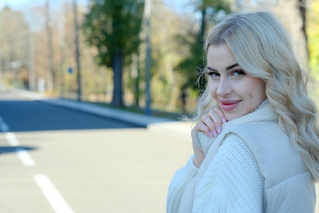 Ritratto di una giovane bellissima bionda con i capelli lunghi in un maglione bianco e giacca in una soleggiata passeggiata d'autunno in città