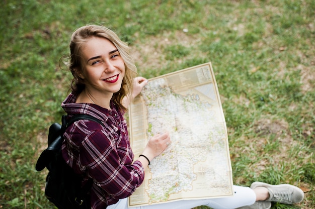 Ritratto di una giovane bionda splendida positiva che si siede sulla terra con una mappa in sue mani nella foresta.