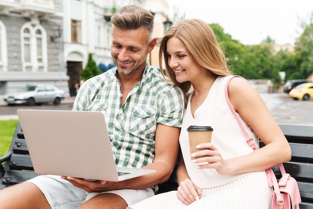 Ritratto di una giovane coppia attraente in abiti estivi che beve caffè e usa il computer portatile mentre è seduto su una panchina in una strada cittadina