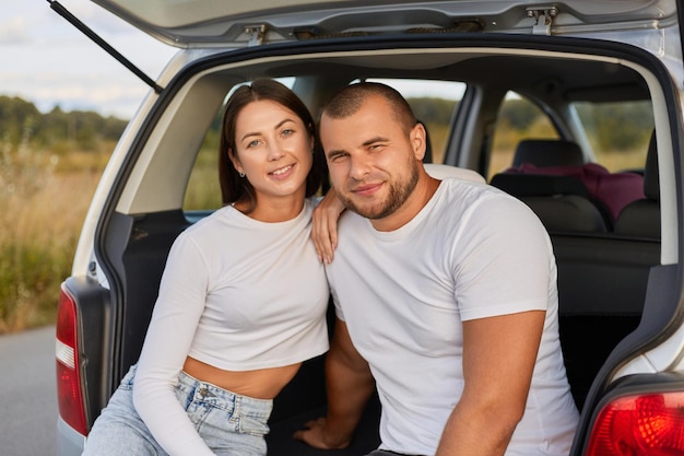 Ritratto di una giovane coppia felice seduta sul bagagliaio dell'auto e guardando la fotocamera con un'espressione facciale positiva soddisfatta che ha viaggiato insieme viaggiando in auto essendo felice